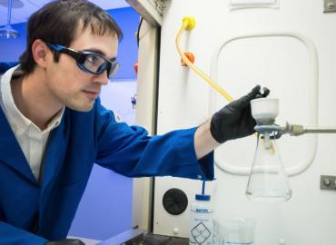 A person in a blue lab coat putting drops in a flask.